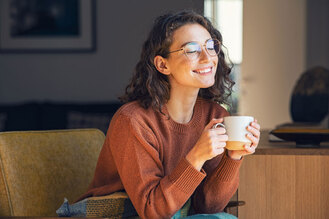 Person with curly hair and glasses wearing rust‑colored sweater, sitting indoors holding white mug with both hands, blurred interior background.